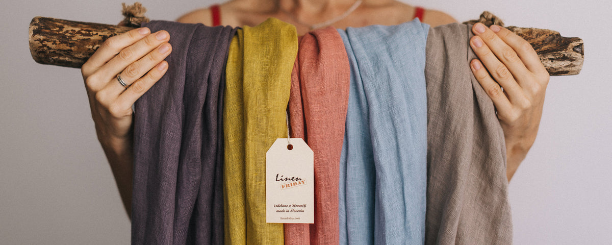 Women hold a thick wooden shelf on which linen scarves of various pastel colors hang.